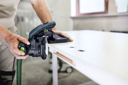 Person sanding the edge of a white wooden board with a handheld electric sander in a workshop.